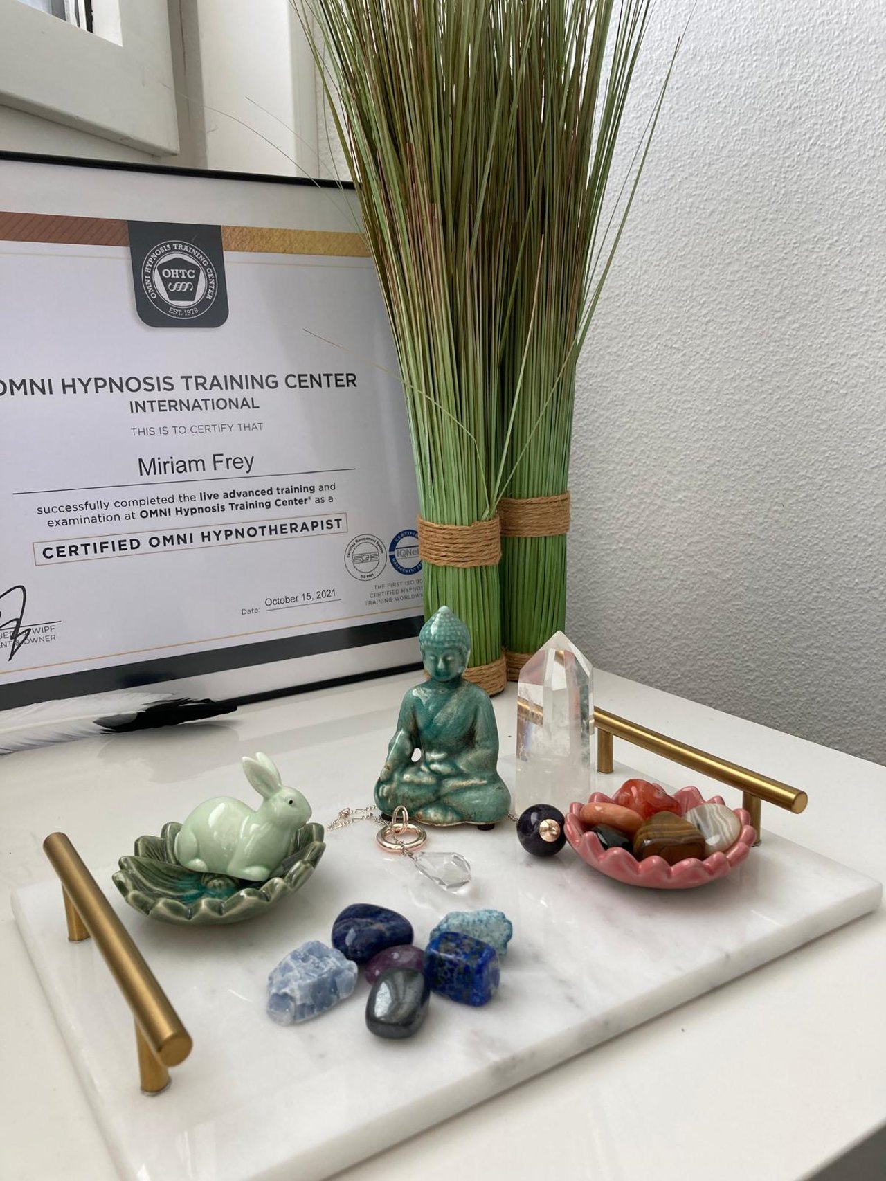 Omni Hypnosis Zertifikat von Miriam Frey neben Buddha-Figur, Heilsteinen und Kristallen auf einem Marmortablett.