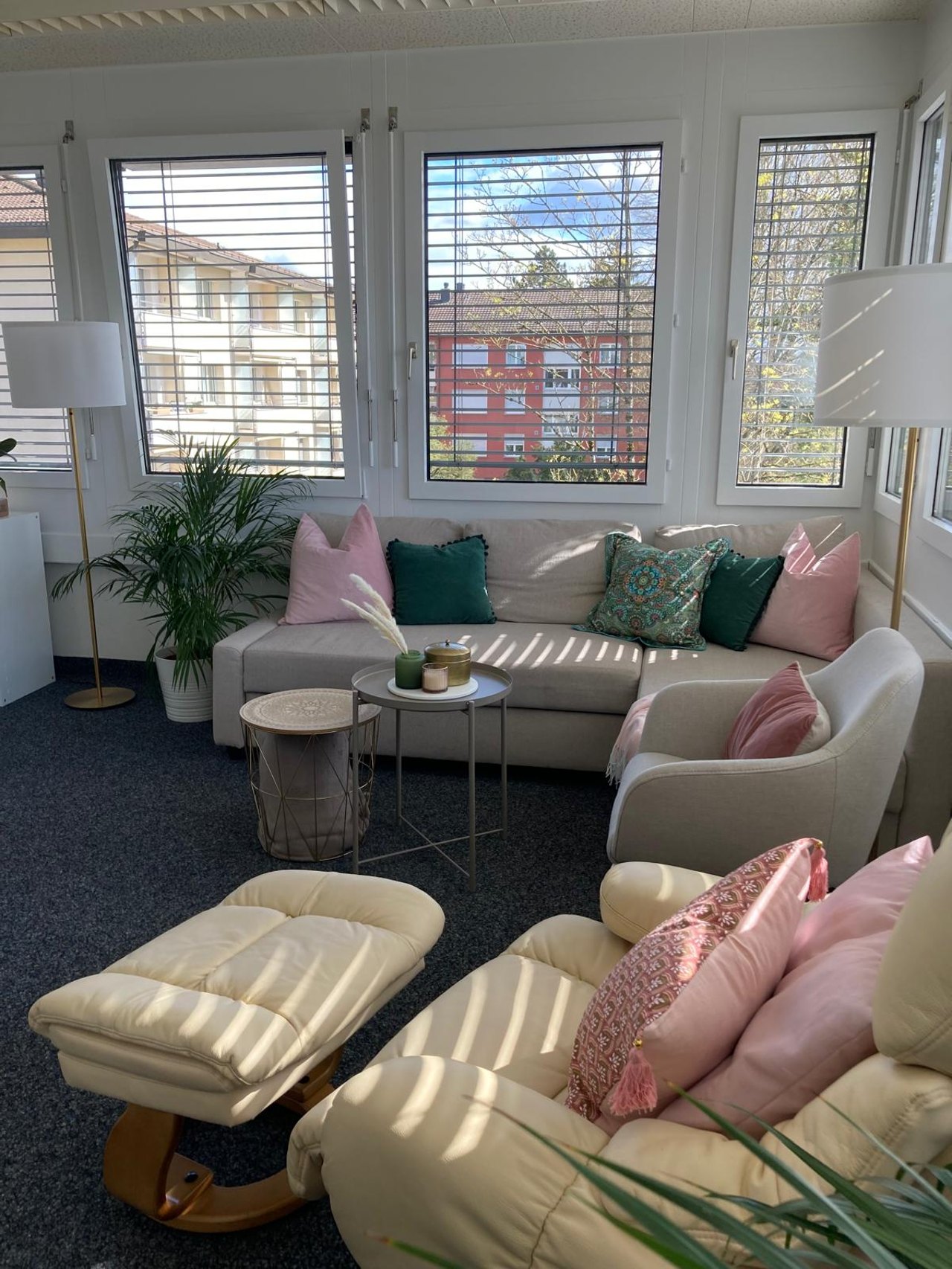 Helles Wohnzimmer mit beigem Sofa, Sesseln, rosa Kissen, Couchtischen und großen Fenstern mit Jalousien.