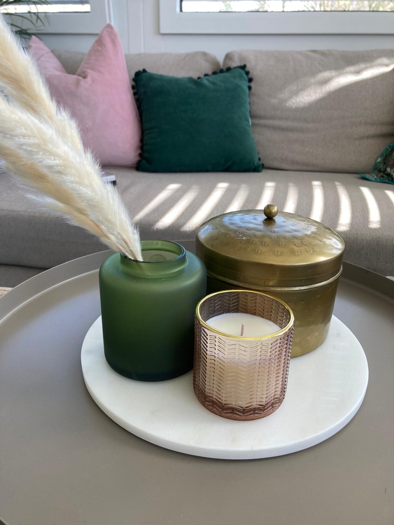 Grüne Vase mit Pampasgras, eine Duftkerze und eine goldene Dose stehen auf einem weißen Marmortablett vor einem Sofa.