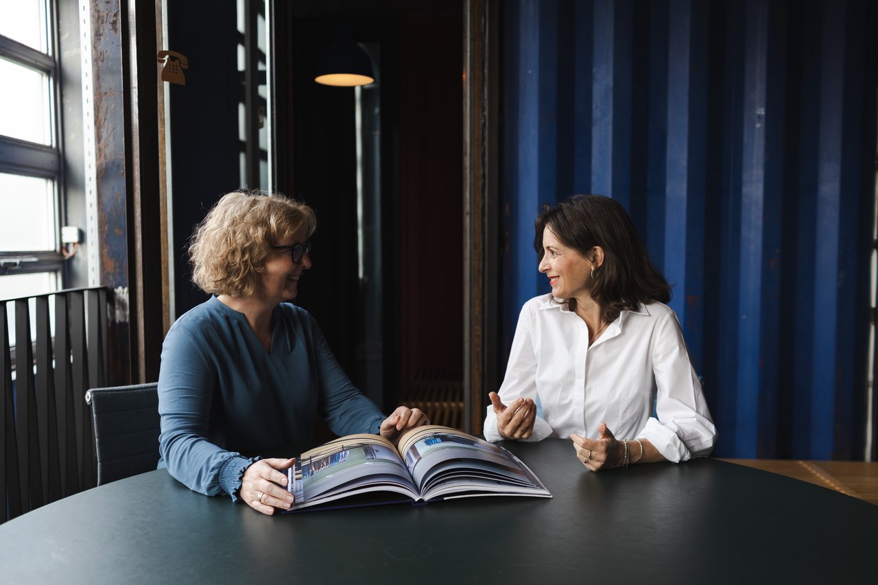 Zwei Frauen sitzen an einem runden Tisch und betrachten gemeinsam ein aufgeschlagenes Buch in einem modernem Büro.