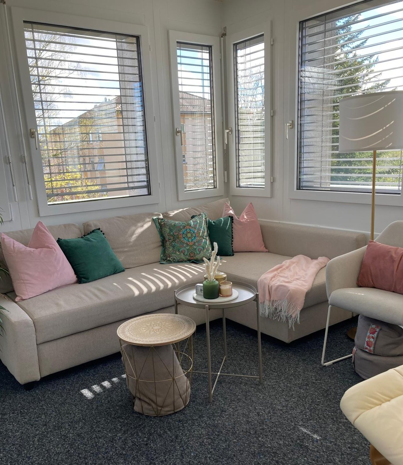 Helles Ecksofa mit rosa und grünen Kissen vor Fenstern mit Jalousien, davor zwei runde Couchtische auf grauem Teppich.